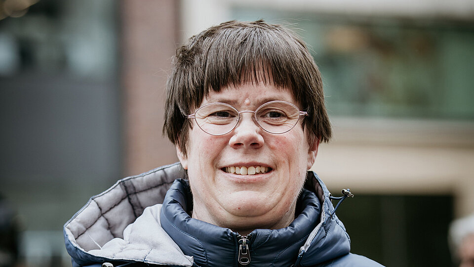 Katrin Schönfeld bei einer Veranstaltung im Freien. Sie trägt eine wattierte, dunkelblaue Jacke mit Kapuze und eine randlose Brille. Sie lächelt in die Kamera und hält ein Schild oder Papier in der Hand. Der Hintergrund ist unscharf, es sind Gebäude und Menschen im öffentlichen Raum zu erkennen.