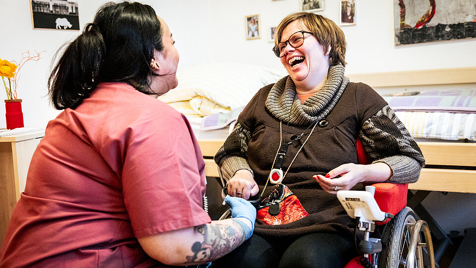 Eine Frau im Rollstuhl und eine Assistenzkraft in roter Arbeitskleidung sitzen einander zugewandt und lachen herzlich miteinander. Die Atmosphäre ist warm und vertraut. Im Hintergrund sind persönliche Gegenstände und Fotos zu sehen, die eine wohnliche Umgebung schaffen.