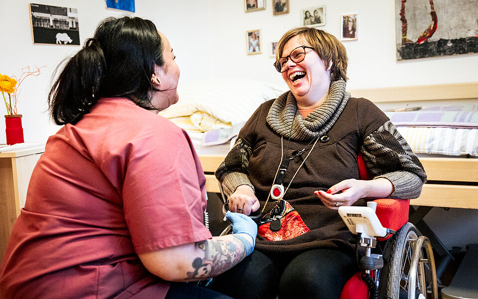 Eine Frau im Rollstuhl und eine Assistenzkraft in roter Arbeitskleidung sitzen einander zugewandt und lachen herzlich miteinander. Die Atmosphäre ist warm und vertraut. Im Hintergrund sind persönliche Gegenstände und Fotos zu sehen, die eine wohnliche Umgebung schaffen.