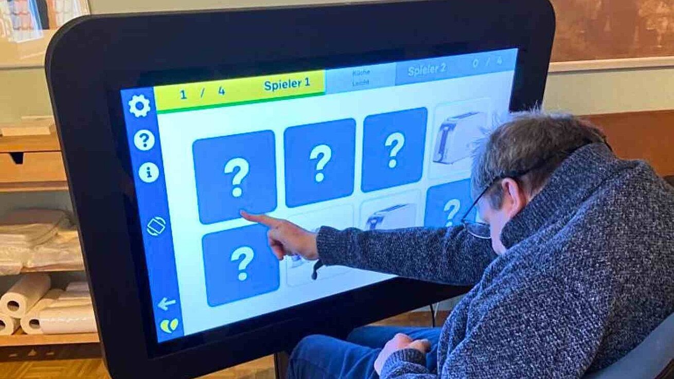 Ein Mensch mit Behinderung sitzt vor einem großen Bildschirm und spielt darauf Memory