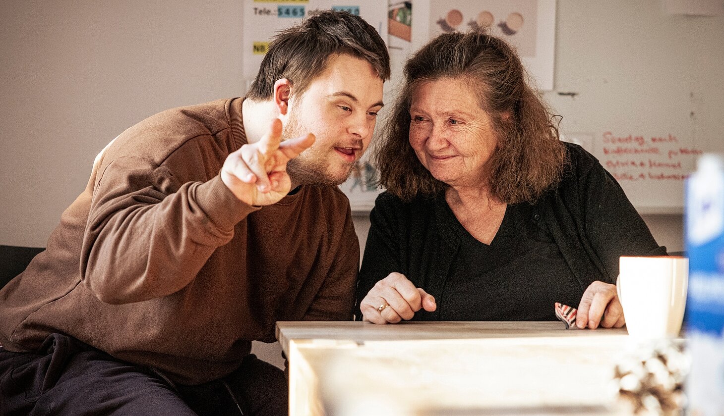 Ein Mann mit Behinderung und eine Assistenzkraft sitzen an einem Tisch eng zusammen. Der Mann macht mit den Fingern das Victory-Zeichen in Richtung Kamera.  