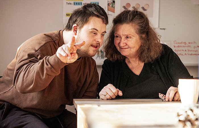 Ein Mann mit Behinderung und eine Assistenzkraft sitzen an einem Tisch eng zusammen. Der Mann macht mit den Fingern das Victory-Zeichen in Richtung Kamera.  
