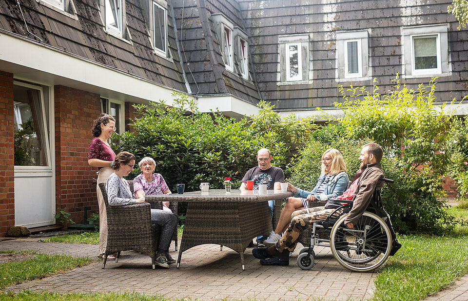 Eine Gruppe von Menschen, darunter mehrere Rollstuhlnutzer*innen, sitzt an einem Tisch im begrünten Innenhof eines Wohnhauses. Eine Betreuungsperson steht daneben.