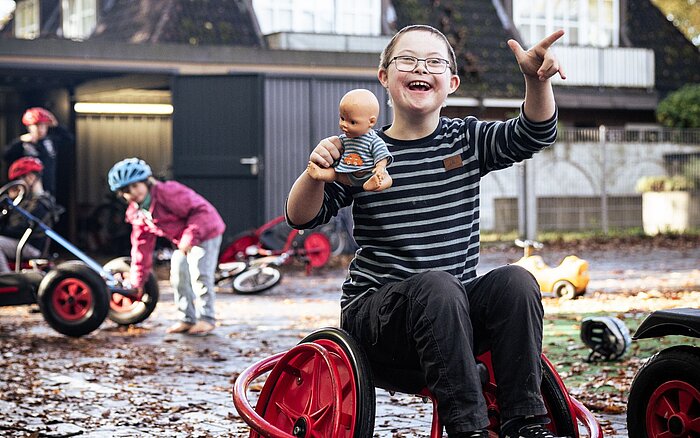 Förderschüler sitzt lachend auf einem Spielgerät im Freien und hält eine Puppe in der Hand, umgeben von anderen Kindern mit Fahrzeugen.