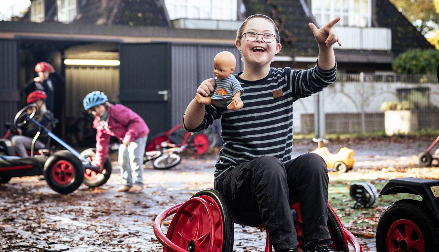 Förderschüler sitzt lachend auf einem Spielgerät im Freien und hält eine Puppe in der Hand, umgeben von anderen Kindern mit Fahrzeugen.