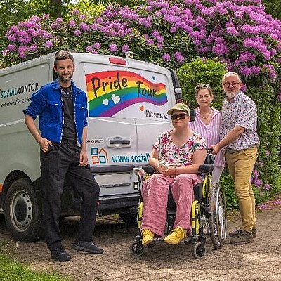 Vier Personen stehen und sitzen neben einem weißen „Pride Tour“-Van mit Regenbogenmotiv. Eine Person im Rollstuhl sitzt vorne, drei weitere Personen stehen daneben, darunter eine Person im blauen Hemd und eine Person mit Brille. Im Hintergrund blühende rosa Sträucher und Bäume in einer grünen Park- oder Wohnumgebung.