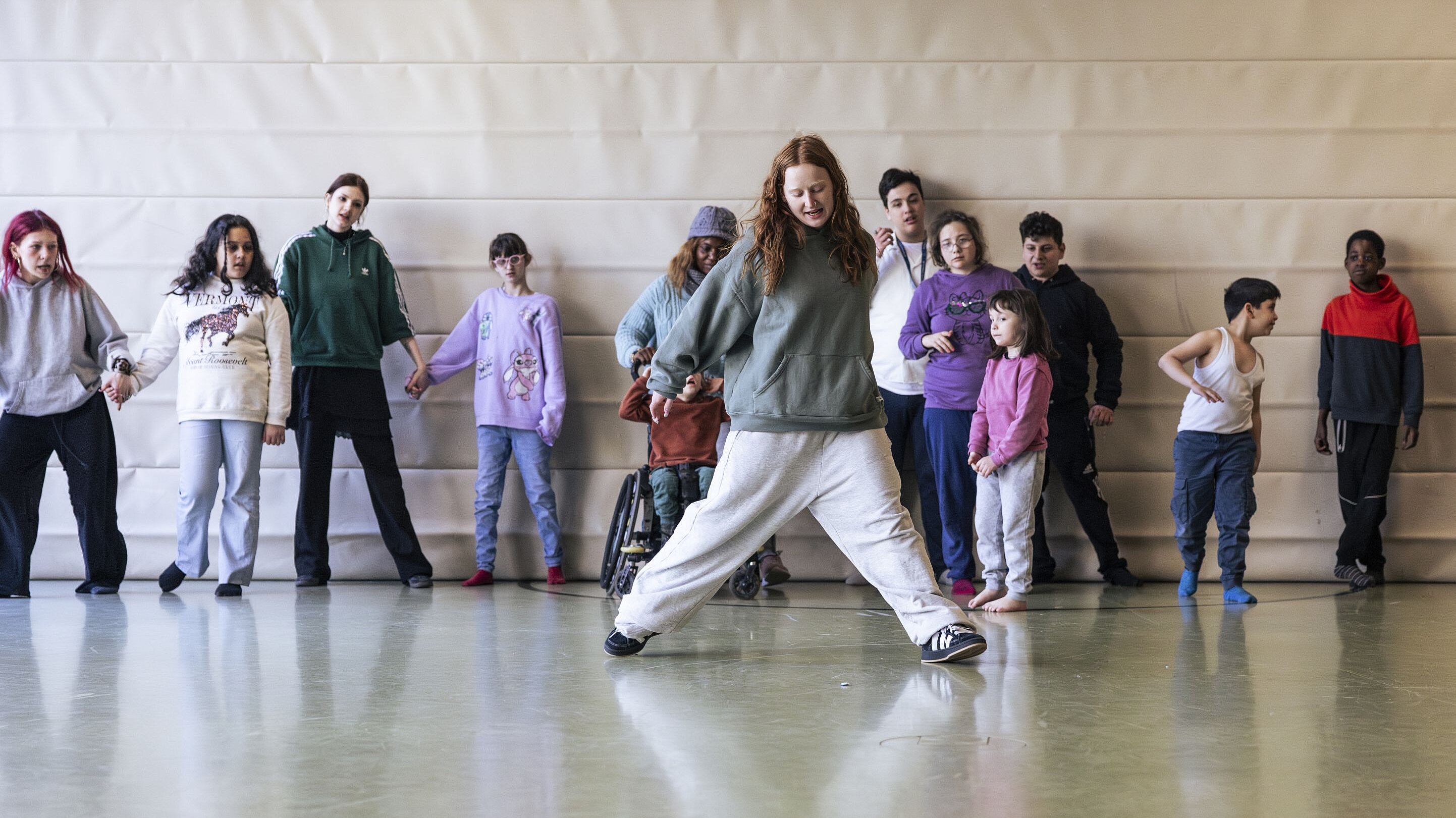Eine Gruppe von Kindern und Jugendlichen steht in einer Reihe, hält sich an den Händen und beobachtet eine Tänzerin, die eine dynamische Hip-Hop-Bewegung vormacht. Die Szene spielt sich in einer Turnhalle ab und zeigt gemeinschaftliches Lernen und Bewegung.