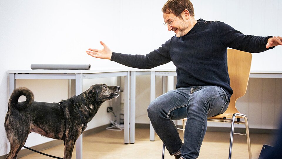 Anleiter begrüßt Hund im Gruppenraum, Hund steht vor ihm, Arme weit geöffnet.