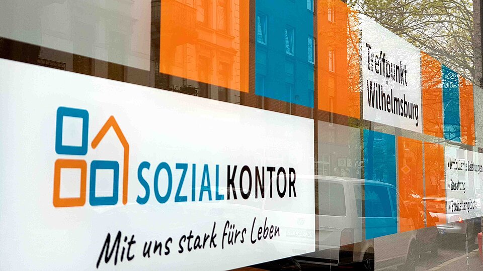 Schaufensterfront des Sozialkontors mit dem Logo und dem Schriftzug „Mit uns stark fürs Leben“. Auf der Glasfläche sind farbige Folien in Orange und Blau sowie die Beschriftung „Treffpunkt Wilhelmsburg“ zu sehen.