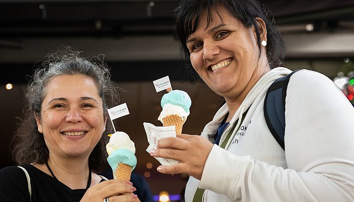 Ilkay Boran und Sarah Bartsch stehen lächelnd nebeneinander und halten Eiswaffeln mit blau-weißem Eis in den Händen.
