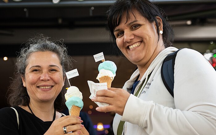 Ilkay Boran und Sarah Bartsch stehen lächelnd nebeneinander und halten Eiswaffeln mit blau-weißem Eis in den Händen.
