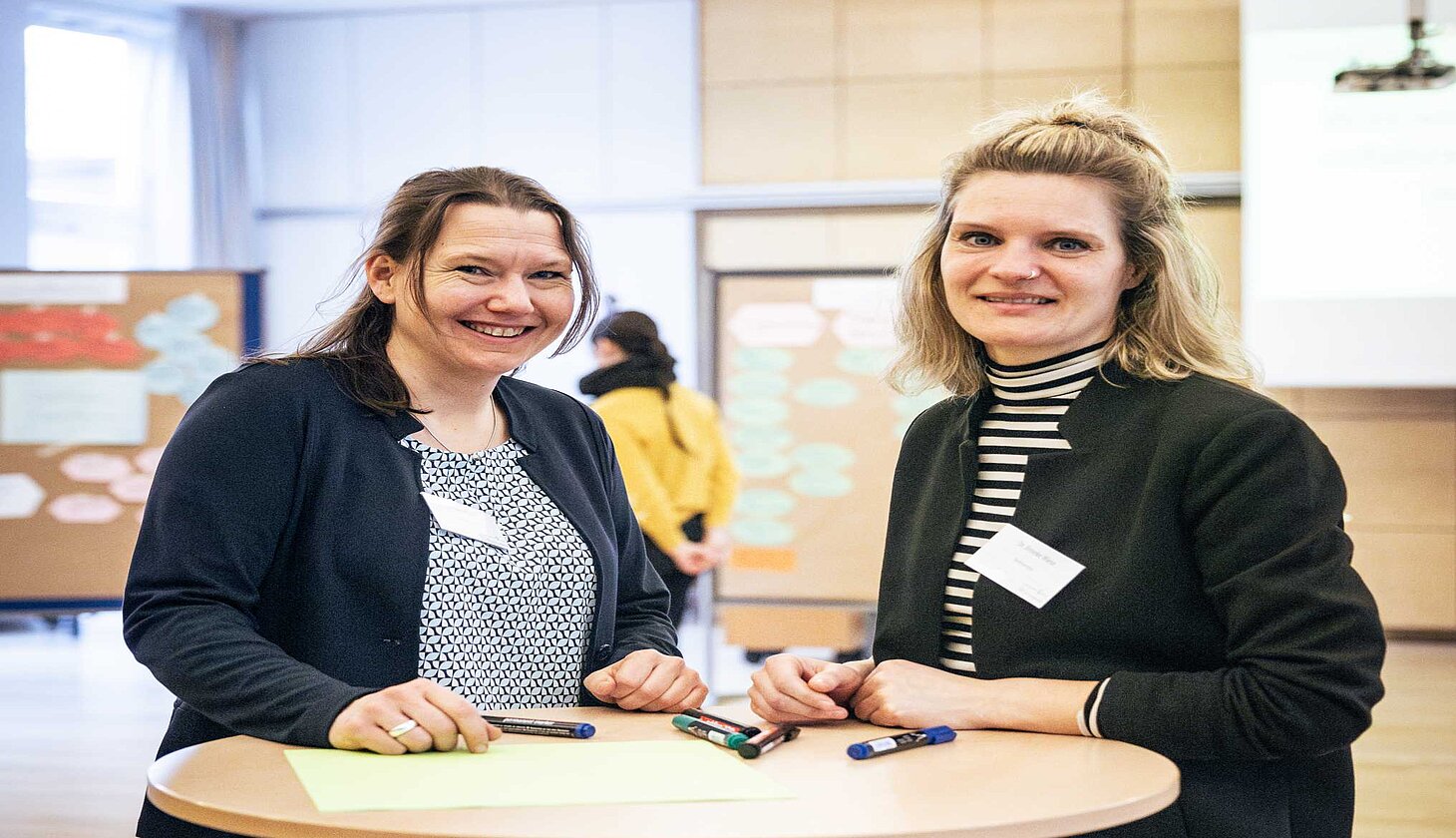 Zwei Frauen stehen an einem Stehtisch mit Markern und Papier und blicken lächelnd in die Kamera. Im Hintergrund ist der Workshopraum mit beschrifteten und beklebten Pinnwänden zu sehen.