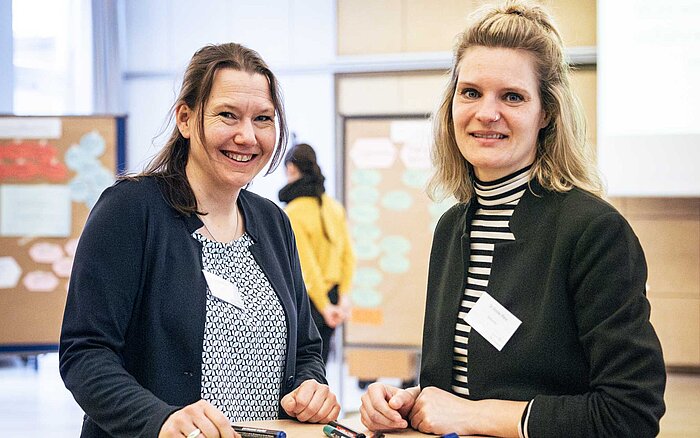 Zwei Frauen stehen an einem Stehtisch mit Markern und Papier und blicken lächelnd in die Kamera. Im Hintergrund ist der Workshopraum mit beschrifteten und beklebten Pinnwänden zu sehen.