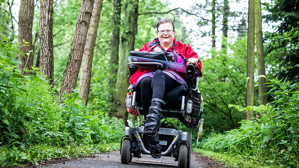 Eine Frau im Rollstuhl fährt auf einem Waldweg. Sie trägt eine rote Jacke und lächelt in die Kamera. Die Umgebung ist grün und dicht bewaldet.