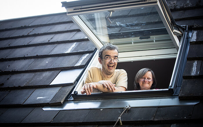 Zwei Nutzer*innen des Sozialkontors schauen fröhlich aus einem geöffneten Dachfenster. Der Mann vorne trägt eine Brille und ein beiges T-Shirt, die Frau hinter ihm lächelt und hat schulterlanges Haar. Das Dach ist mit dunklen Schindeln gedeckt.