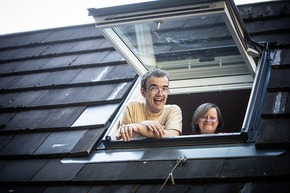 Zwei Nutzer*innen des Sozialkontors schauen fröhlich aus einem geöffneten Dachfenster. Der Mann vorne trägt eine Brille und ein beiges T-Shirt, die Frau hinter ihm lächelt und hat schulterlanges Haar. Das Dach ist mit dunklen Schindeln gedeckt.