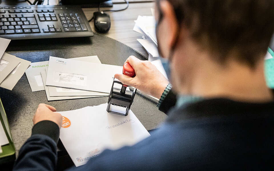Eine Person sitzt am Schreibtisch und stempelt Dokumente. Auf dem Tisch liegen Papierstapel und ein Computer ist im Hintergrund sichtbar. 