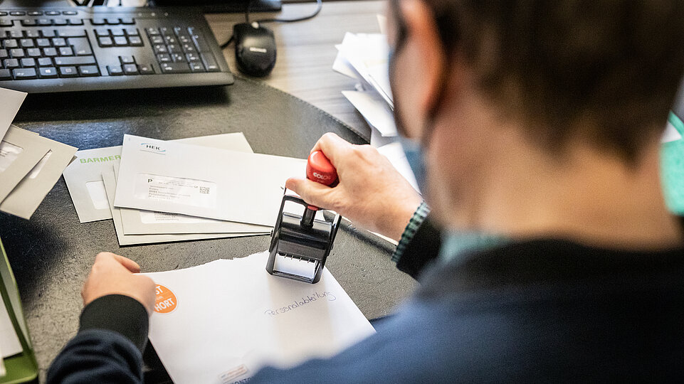 Eine Person sitzt am Schreibtisch und stempelt Dokumente. Auf dem Tisch liegen Papierstapel und ein Computer ist im Hintergrund sichtbar.