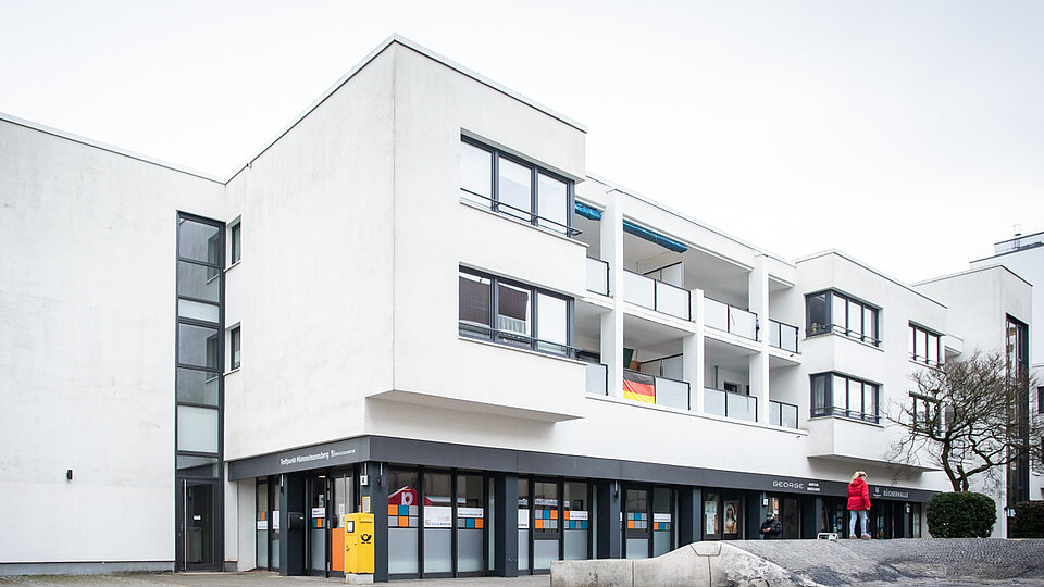 Mehrgeschossiges Wohn- und Geschäftshaus mit weißen Fassaden, Balkonen und großen Fensterflächen. Im Erdgeschoss befinden sich Laden- oder Büroräume, davor verläuft ein gepflasterter Platz.
