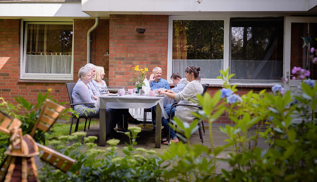 Mehrere Personen sitzen an einem gedeckten Tisch auf einer Terrasse vor einem Backsteingebäude. Im Vordergrund sind Gartenpflanzen zu sehen, auf dem Tisch stehen Getränke und Blumen.