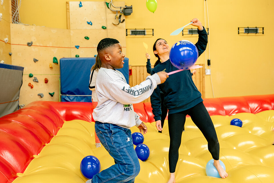 Ein Junge und eine junge Erwachsene balancieren auf einem großen gelben Luftkissen und spielen mit Luftballons in einer Turnhalle, während andere Kinder im Hintergrund aktiv sind.