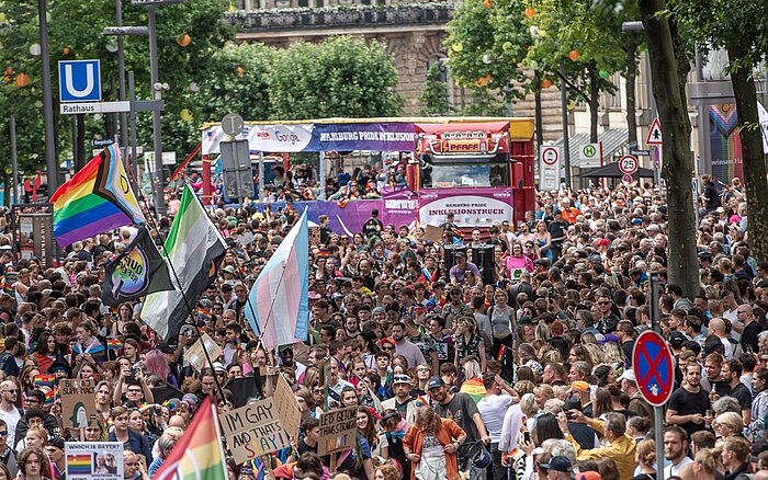 Dichte Menschenmenge bei der CSD-Demonstration in Hamburg. Im Hintergrund fährt der violett-rote Hamburg Pride Inklusionstruck, im Vordergrund wehen verschiedene Pride-Flaggen.