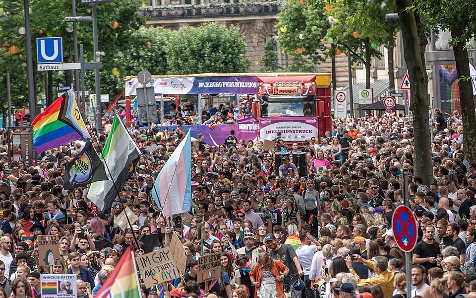 Dichte Menschenmenge bei der CSD-Demonstration in Hamburg. Im Hintergrund fährt der violett-rote Hamburg Pride Inklusionstruck, im Vordergrund wehen verschiedene Pride-Flaggen.
