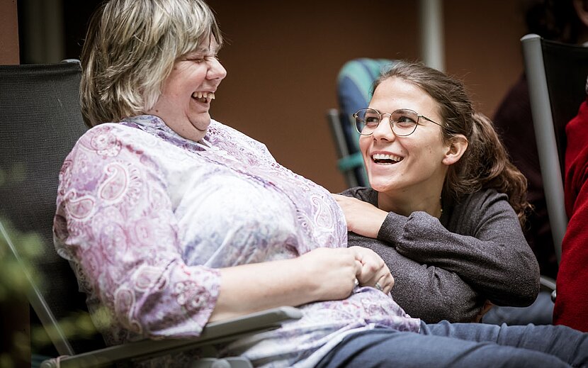 Zwei Frauen lachen herzlich miteinander. Eine von ihnen sitzt im Rollstuhl, die andere lehnt sich lachend zu ihr.
