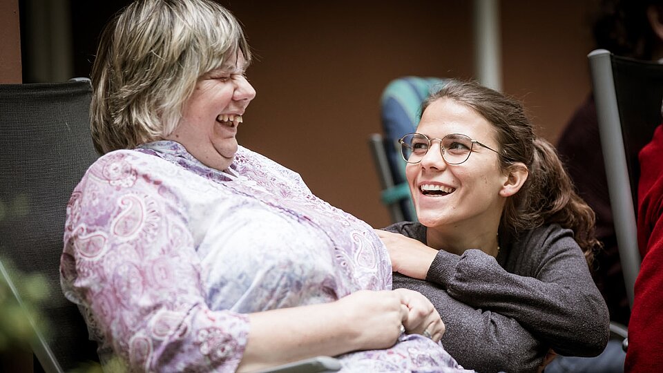 Zwei Frauen lachen herzlich miteinander. Eine von ihnen sitzt im Rollstuhl, die andere lehnt sich lachend zu ihr.
