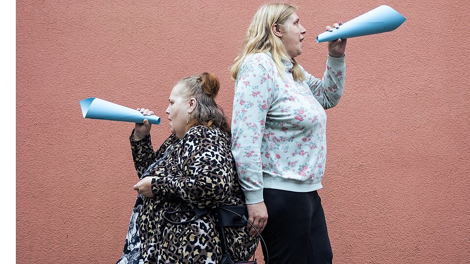 Zwei Frauen Rücken an Rücken, eine sitzt auf einem Rollator. In den Händen halten sie jeweils ein wie ein Sprachrohr geformten Papier durch das sie sprechen.
