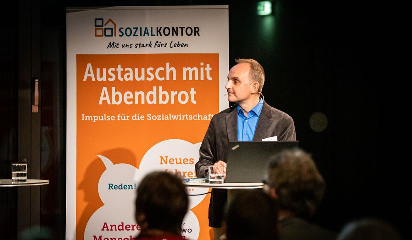 Eine Person steht an einem Stehtisch mit Laptop vor einem Roll-up des Sozialkontors. Auf dem Banner steht unter anderem „Austausch mit Abendbrot – Impulse für die Sozialwirtschaft“, im Vordergrund sind Zuhörende zu sehen.
