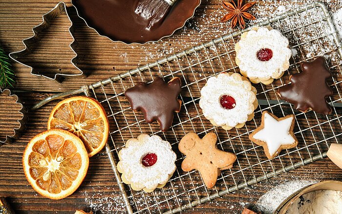 Verschiedene weihnachtliche Kekse, darunter mit Schokolade überzogene, mit Puderzucker bestreute und sternförmige, liegen auf einem Gitterrost. Drumherum liegen getrocknete Orangenscheiben, Ausstechformen und Tannenzweige auf einer Holzoberfläche.