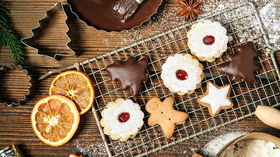 Verschiedene weihnachtliche Kekse, darunter mit Schokolade überzogene, mit Puderzucker bestreute und sternförmige, liegen auf einem Gitterrost. Drumherum liegen getrocknete Orangenscheiben, Ausstechformen und Tannenzweige auf einer Holzoberfläche.