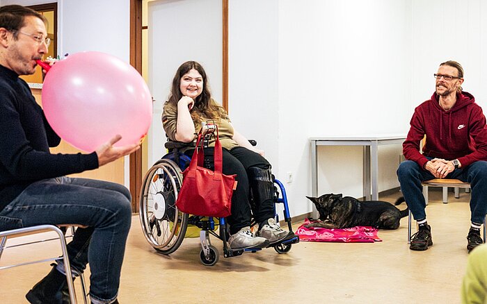 Bewegungsgruppe drinnen: Mann bläst Ballon auf, Rollstuhlnutzerin und weiterer Teilnehmer lachen, Hund liegt dabei.