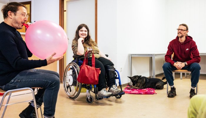 Bewegungsgruppe drinnen: Mann bläst Ballon auf, Rollstuhlnutzerin und weiterer Teilnehmer lachen, Hund liegt dabei.