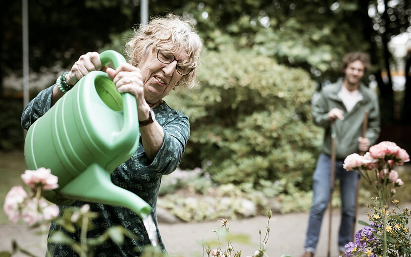 Eine ältere Frau mit Brille gießt mit einer großen grünen Gießkanne Blumen in einem Garten. Im Hintergrund steht ein Mann mit einem Gartenwerkzeug und schaut ihr lächelnd zu.