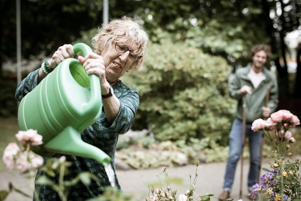 Eine ältere Frau mit Brille gießt mit einer großen grünen Gießkanne Blumen in einem Garten. Im Hintergrund steht ein Mann mit einem Gartenwerkzeug und schaut ihr lächelnd zu.