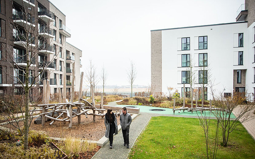 Zwei Personen gehen einen Weg entlang durch einen modernen Wohnkomplex mit barrierefreier Gestaltung. Umgeben sind sie von Hochbeeten, Sitzgelegenheiten und jungen Bäumen. Im Hintergrund sind mehrere mehrstöckige Gebäude mit Balkonen zu sehen.