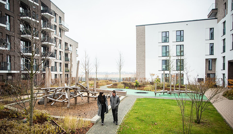 Zwei Personen gehen einen Weg entlang durch einen modernen Wohnkomplex mit barrierefreier Gestaltung. Umgeben sind sie von Hochbeeten, Sitzgelegenheiten und jungen Bäumen. Im Hintergrund sind mehrere mehrstöckige Gebäude mit Balkonen zu sehen.