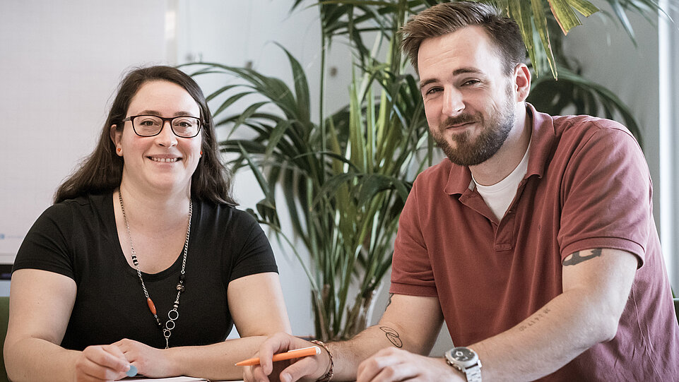 Zwei Personen sitzen nebeneinander an einem Tisch und blicken freundlich in die Kamera. Auf dem Tisch liegen Schreibutensilien. Im Hintergrund stehen große Pflanzen.