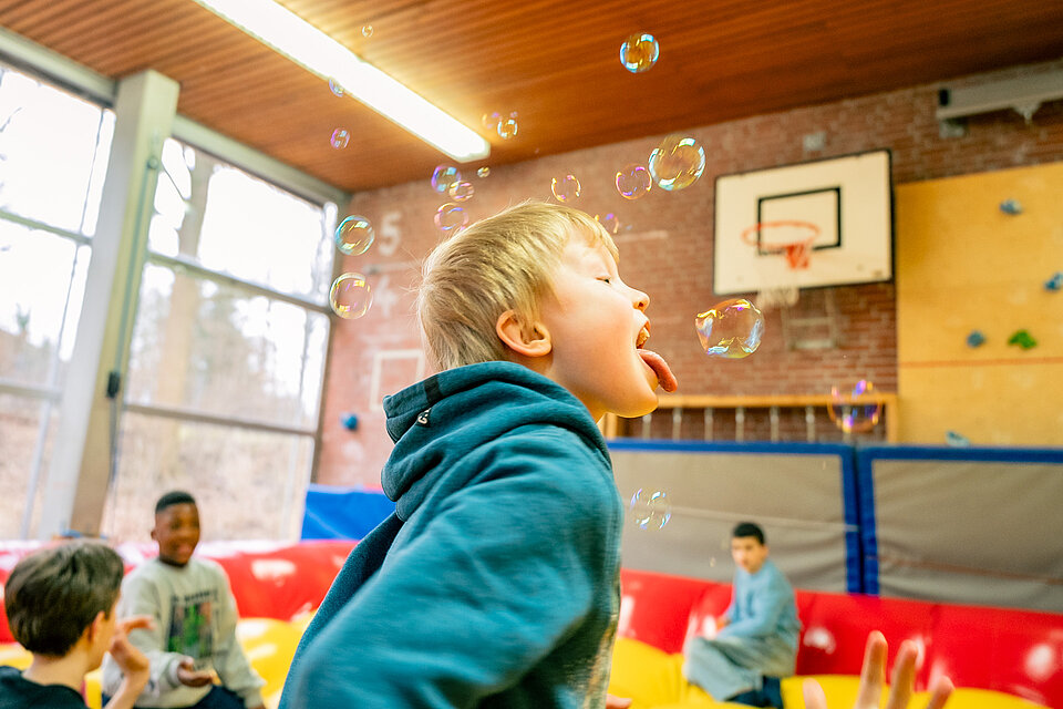 Ein Kind jagt aufgeregt Seifenblasen in einer Turnhalle, mit einem breiten Lächeln im Gesicht, während es die Zunge nach einer schwebenden Seifenblase streckt.