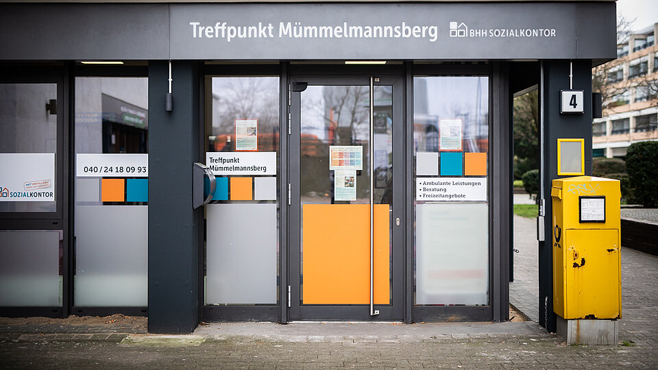 Eingangsbereich des „Treffpunkt Mümmelmannsberg“ des Sozialkontors mit verglaster Tür und Fensterfront. Neben dem Eingang steht ein gelber Briefkasten, über der Tür befindet sich die Beschriftung des Standorts.
