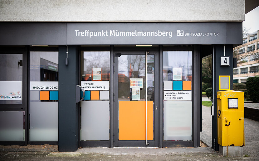 Eingangsbereich des „Treffpunkt Mümmelmannsberg“ des Sozialkontors mit verglaster Tür und Fensterfront. Neben dem Eingang steht ein gelber Briefkasten, über der Tür befindet sich die Beschriftung des Standorts.