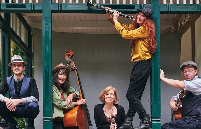 Ein Pressefoto der Band Mischpoke. Die Band posiert entspannt und fröhlich mit ihren unterschiedlichen Instrumenten in einem offenen Pavillon mit grünen Holzbalken. Zwei sitzen an den Seiten, einer hält eine Gitarre, der andere einen Kontrabass. In der Mitte steht eine Frau mit einer Geige. Eine weitere Person steht erhöht und spielt eine Klarinette.