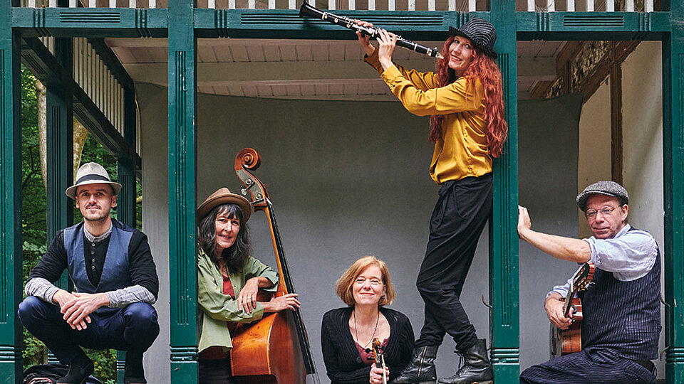 Ein Pressefoto der Band Mischpoke. Die Band posiert entspannt und fröhlich mit ihren unterschiedlichen Instrumenten in einem offenen Pavillon mit grünen Holzbalken. Zwei sitzen an den Seiten, einer hält eine Gitarre, der andere einen Kontrabass. In der Mitte steht eine Frau mit einer Geige. Eine weitere Person steht erhöht und spielt eine Klarinette.