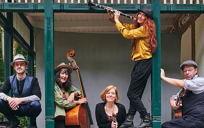 Ein Pressefoto der Band Mischpoke. Die Band posiert entspannt und fröhlich mit ihren unterschiedlichen Instrumenten in einem offenen Pavillon mit grünen Holzbalken. Zwei sitzen an den Seiten, einer hält eine Gitarre, der andere einen Kontrabass. In der Mitte steht eine Frau mit einer Geige. Eine weitere Person steht erhöht und spielt eine Klarinette.