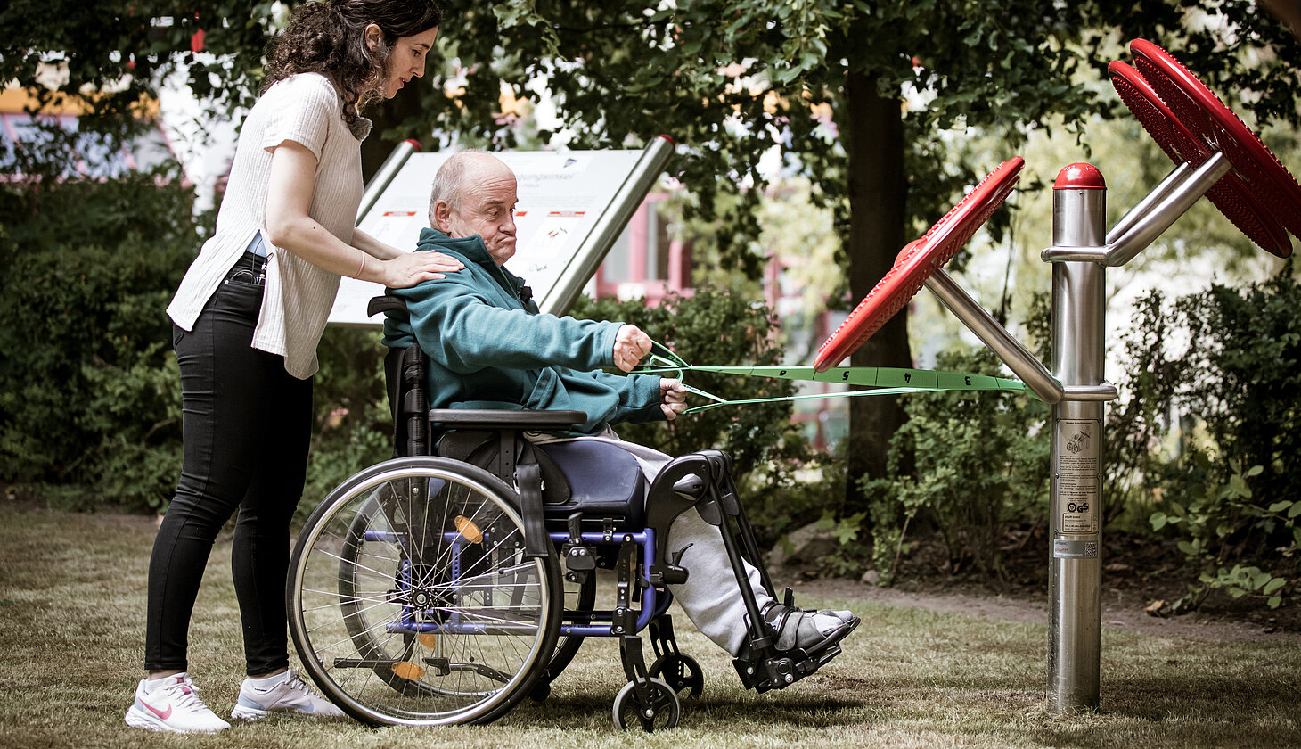 Eine Frau unterstützt einen Mann im Rollstuhl beim Training mit Outdoor-Fitnessgeräten in einem Park bei den 