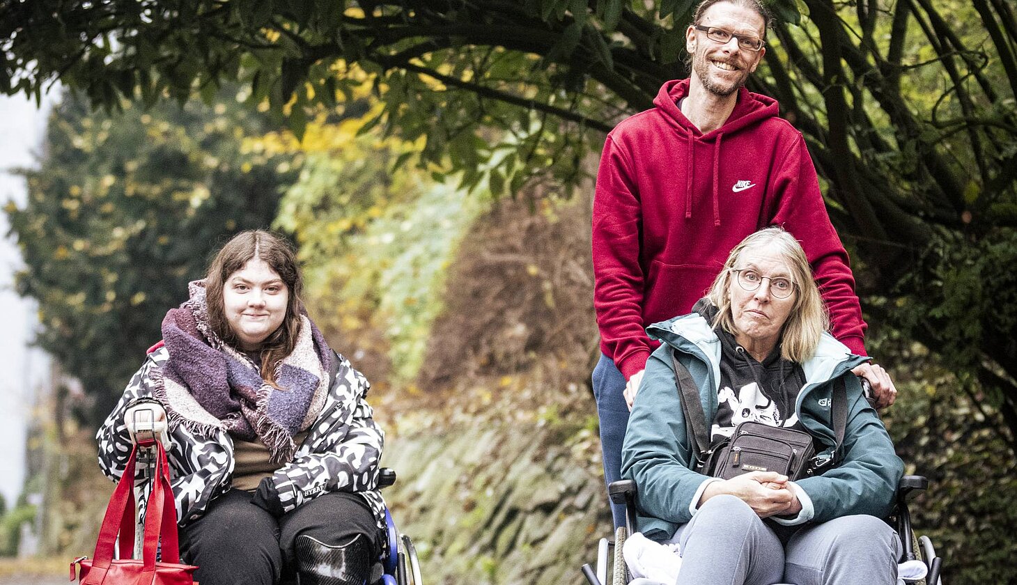 Zwei Frauen im Rollstuhl und ein Mann gehen gemeinsam auf einem herbstlichen Weg unter großen Bäumen entlang.
