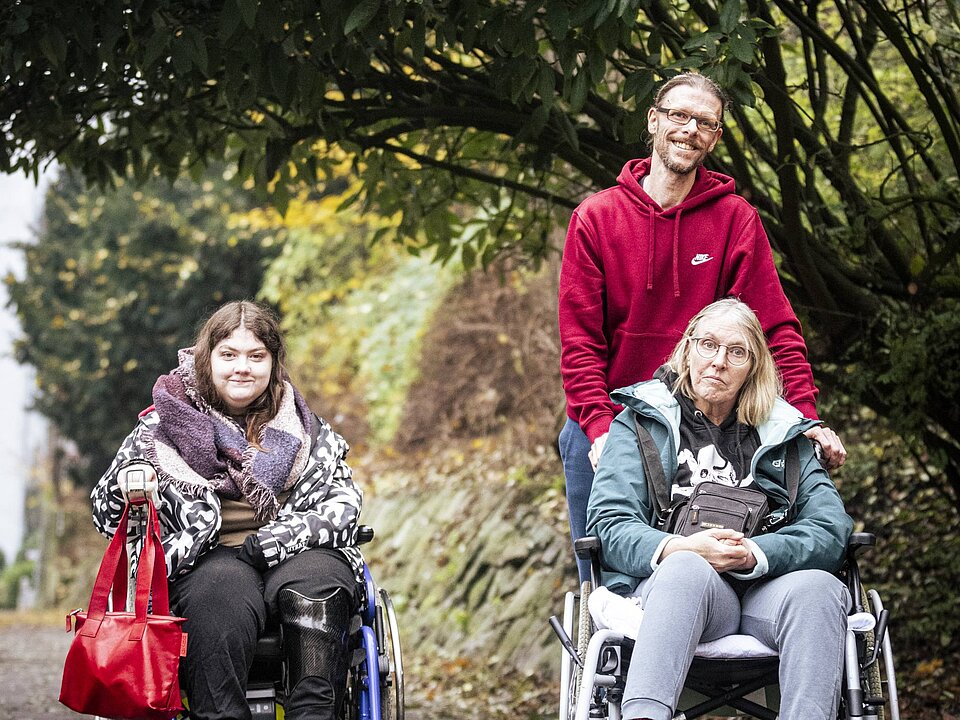 Zwei Frauen im Rollstuhl und ein Mann gehen gemeinsam auf einem herbstlichen Weg unter großen Bäumen entlang.