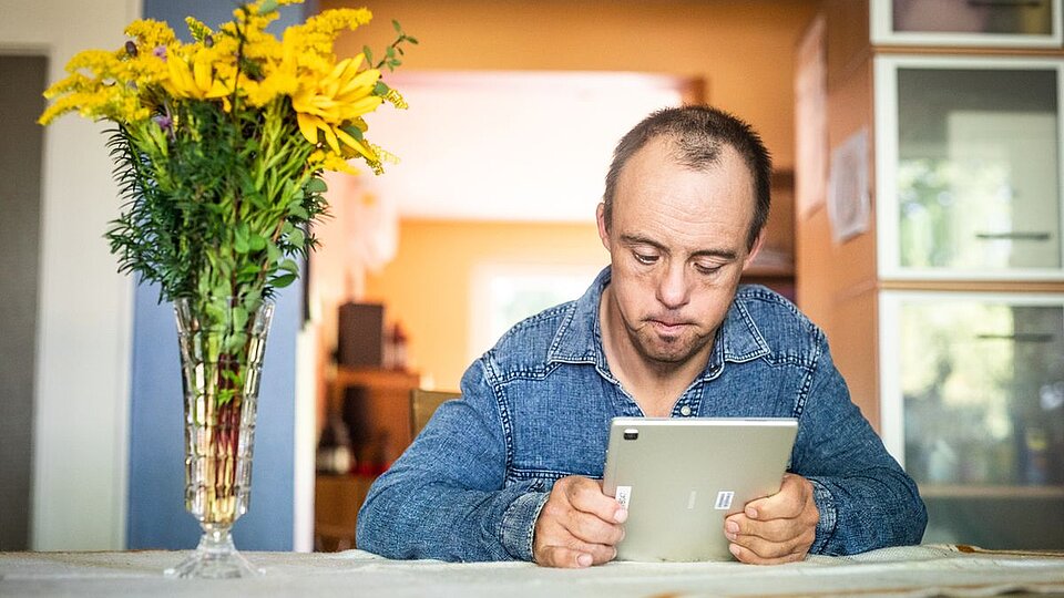 Eine Person sitzt an einem Tisch und hält ein Tablet in beiden Händen. Neben ihr steht eine Vase mit gelben Blumen, im Hintergrund ist ein Wohnraum zu sehen.